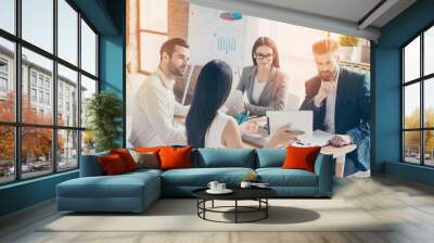 Brunette woman is showing her idea of their company`s development on tablet at the business meeting to her colleagues, all of them are  in formalwear Wall mural